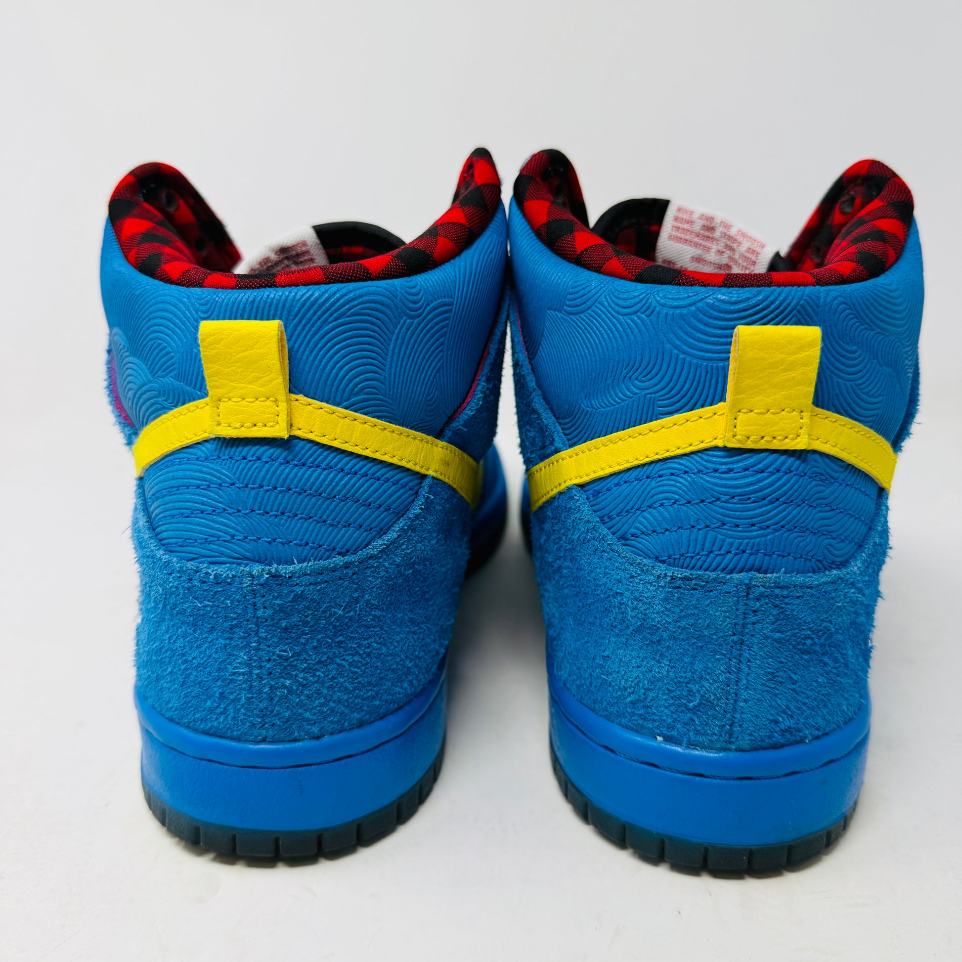 The Nike SB Dunk High Familia Blue Ox, a 2015 release by Nike, features clean blue uppers with a yellow swoosh, decorative stitching, red and black checkered lining, lace-up front, and an edgy skull with crossed bones on the side—its standout design loses no stars.