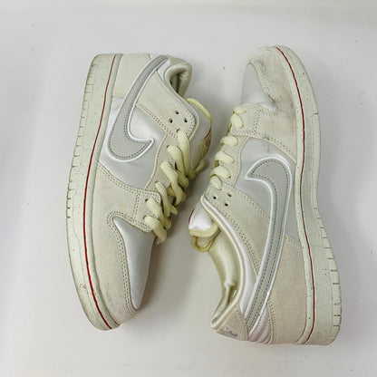 The Nike SB Dunk Low City of Love Light Bone sneakers feature a swoosh logo, off-white color with light cream soles, yellow laces, and red accents around the heel and toe. They have a slightly worn appearance.