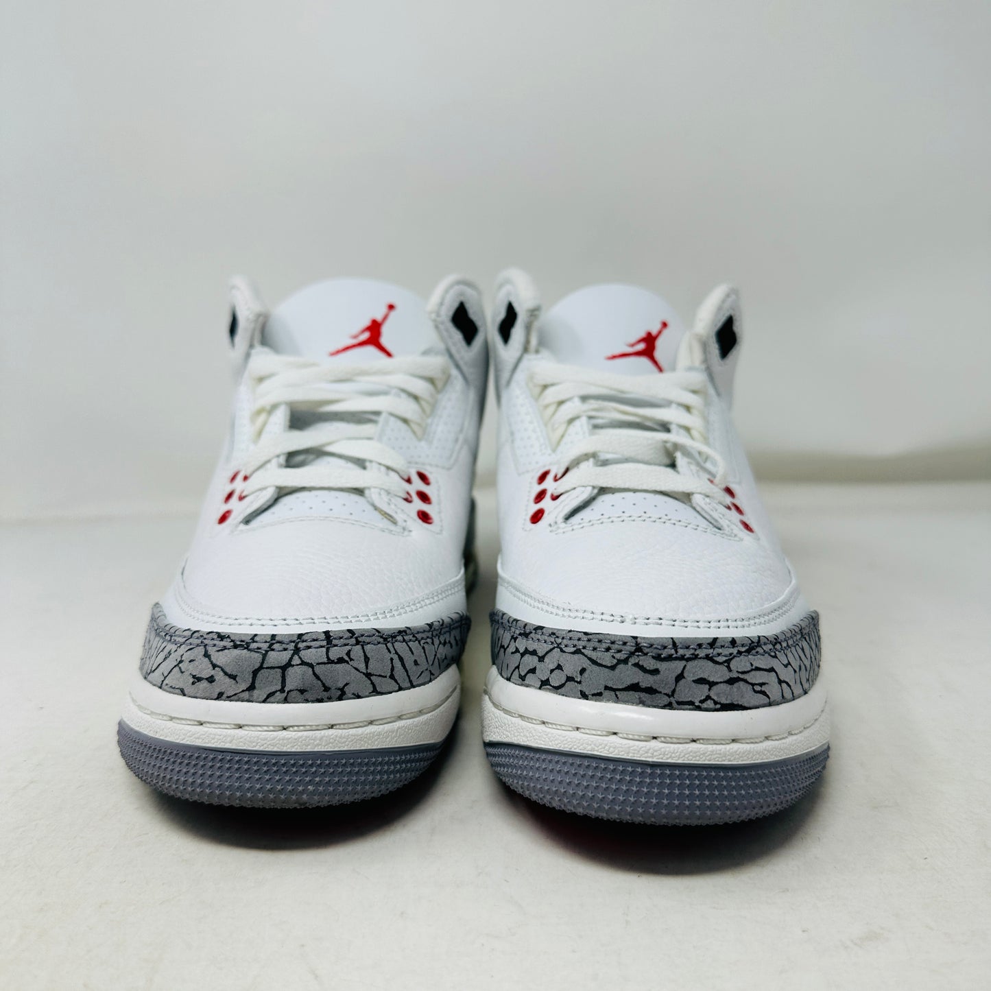 The Jordan 3 Retro White Cement Reimagined (GS) sneakers feature white with gray cracked patterns on toes and sides, red accents on the tongue and eyelets, white laces, and gray soles, photographed against a plain white background.