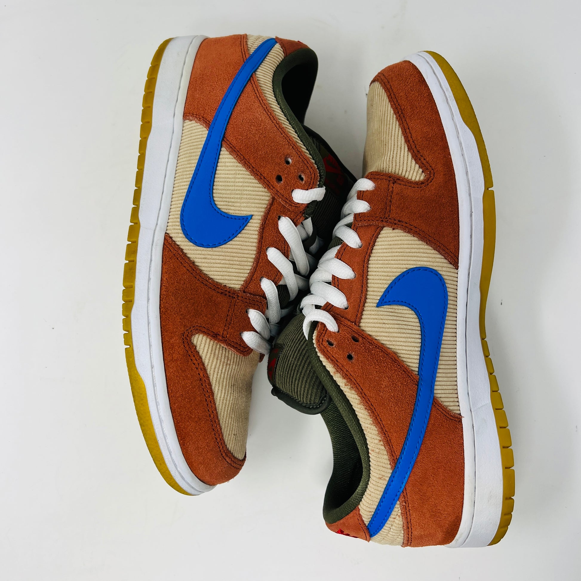 The Nike SB Dunk Low Corduroy Dusty Peach sneakers showcase orange suede with beige corduroy panels and a bright blue swoosh. Olive laces and a yellow sole complete the design, artfully displayed side by side on a white background.