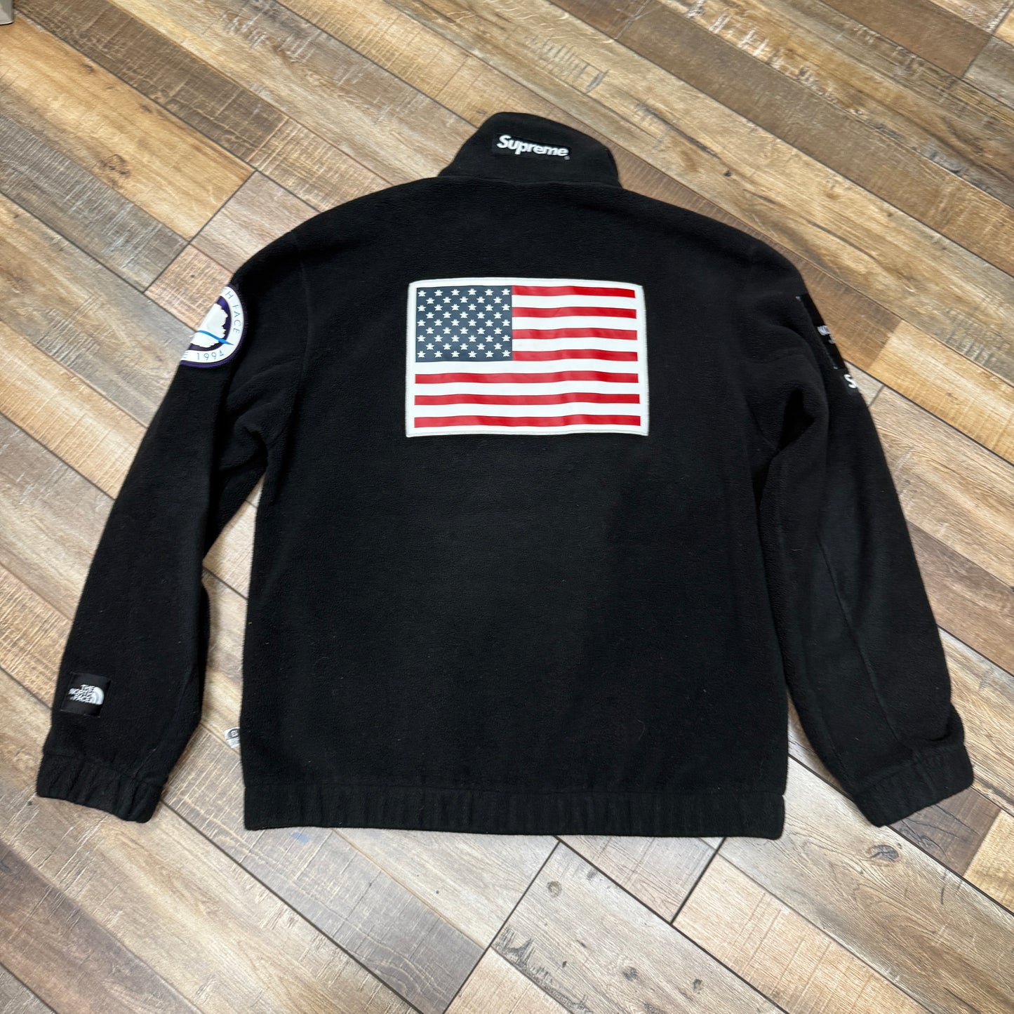 The Supreme The North Face Trans Antarctica Expedition Fleece Jacket in Black showcases The North Face and Supreme logos, a U.S. flag patch on the front, and a circular Antarctica patch on the sleeve. It’s displayed authentically on a wooden floor.