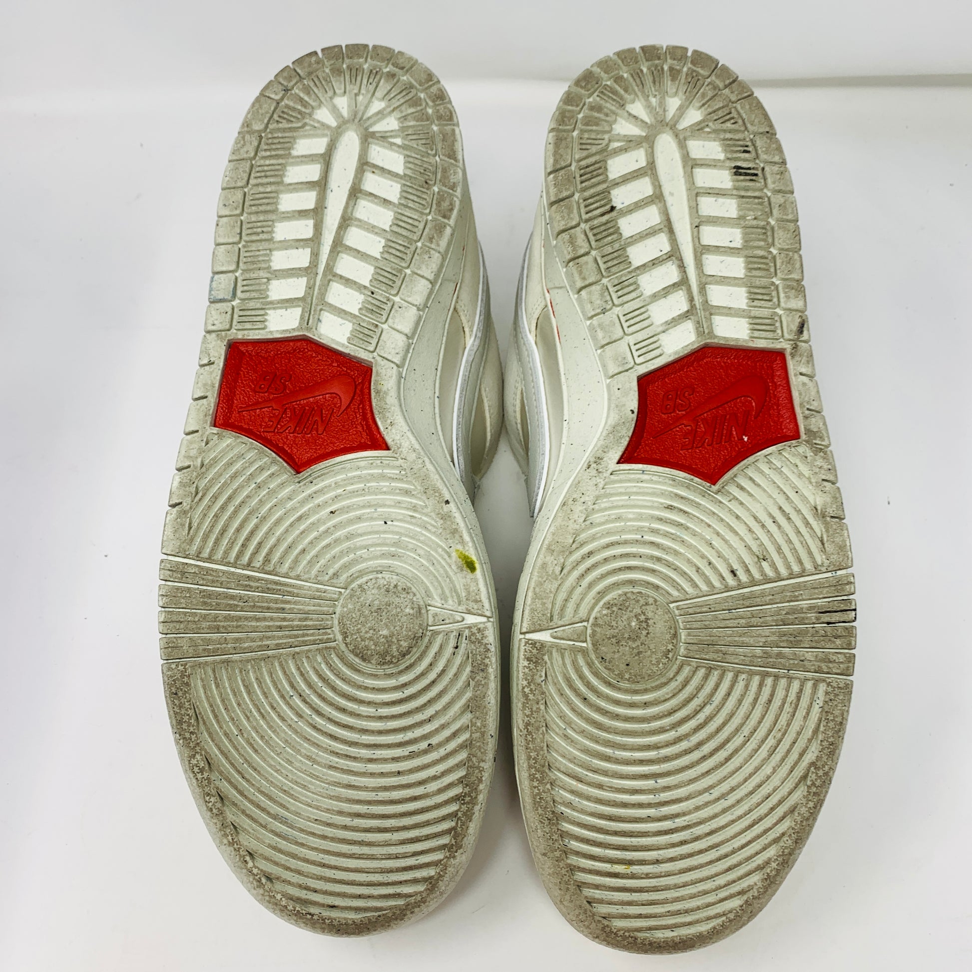 The image features the soles of Nike SB Dunk Low City of Love Light Bone sneakers. The white design includes circular and linear tread patterns with two red hexagonal accents at the ball area displaying Nike. The sneakers are positioned side by side.