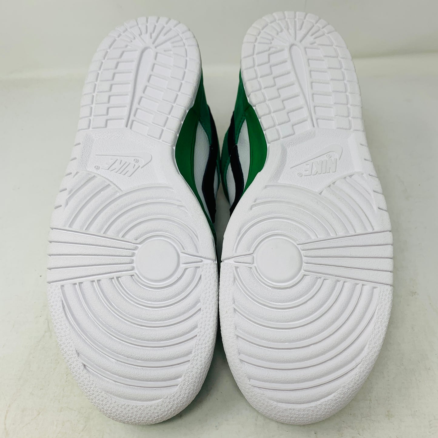 The image displays the soles of Nike SB Dunk Low Heineken athletic shoes, featuring white textured bottoms with circular patterns and grooves, while the midsole and sides are green and white.