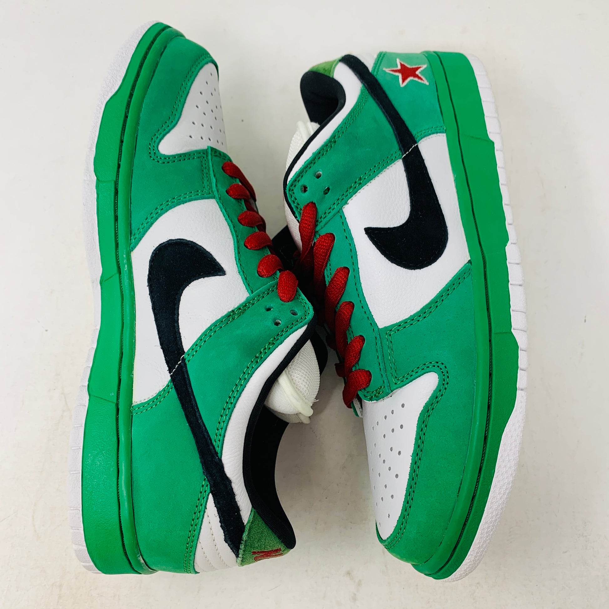 The Nike SB Dunk Low Heineken features green and white sneakers with black swoosh logos, red laces, and a red star detail on the heel against a white background.