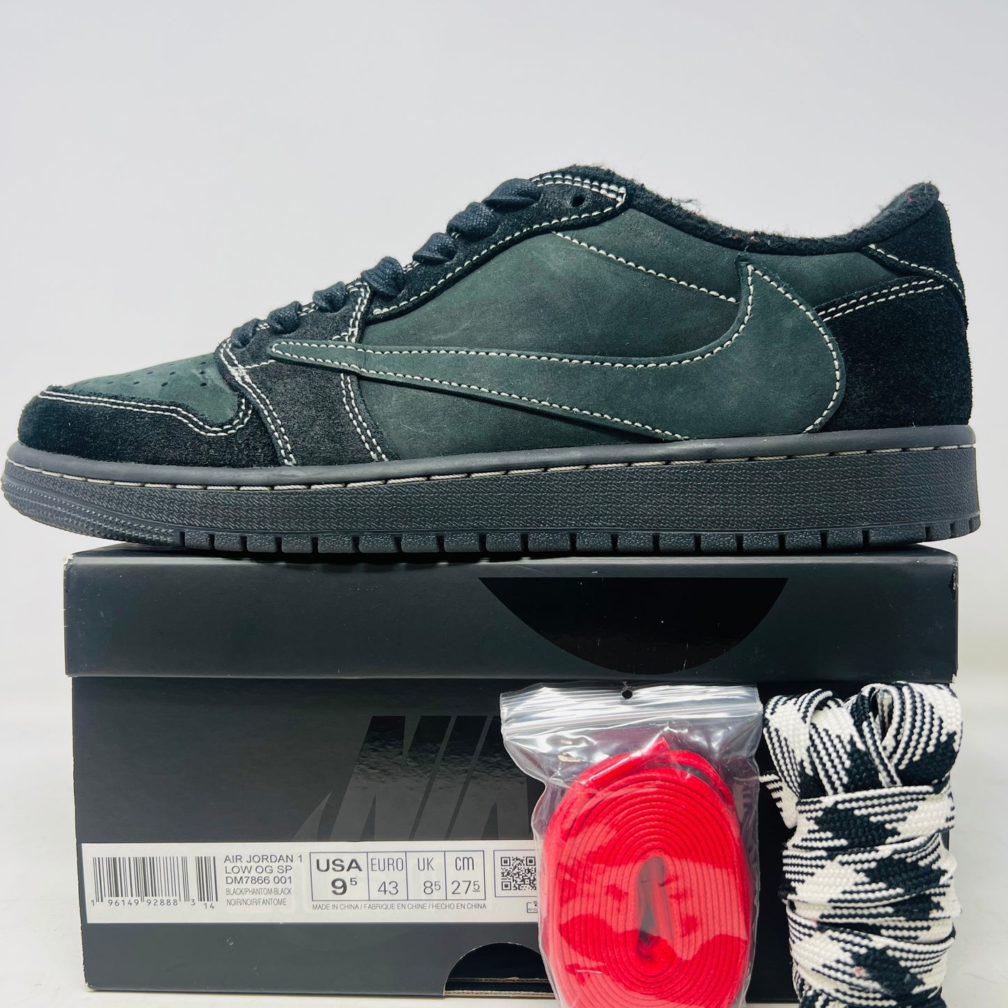 The Jordan 1 Retro Low OG SP Travis Scott Black Phantom is a stylish sneaker featuring a dark green suede upper with black leather accents, packed in a black shoebox. It comes with red and black-and-white patterned spare laces, showcasing its original swoosh design.