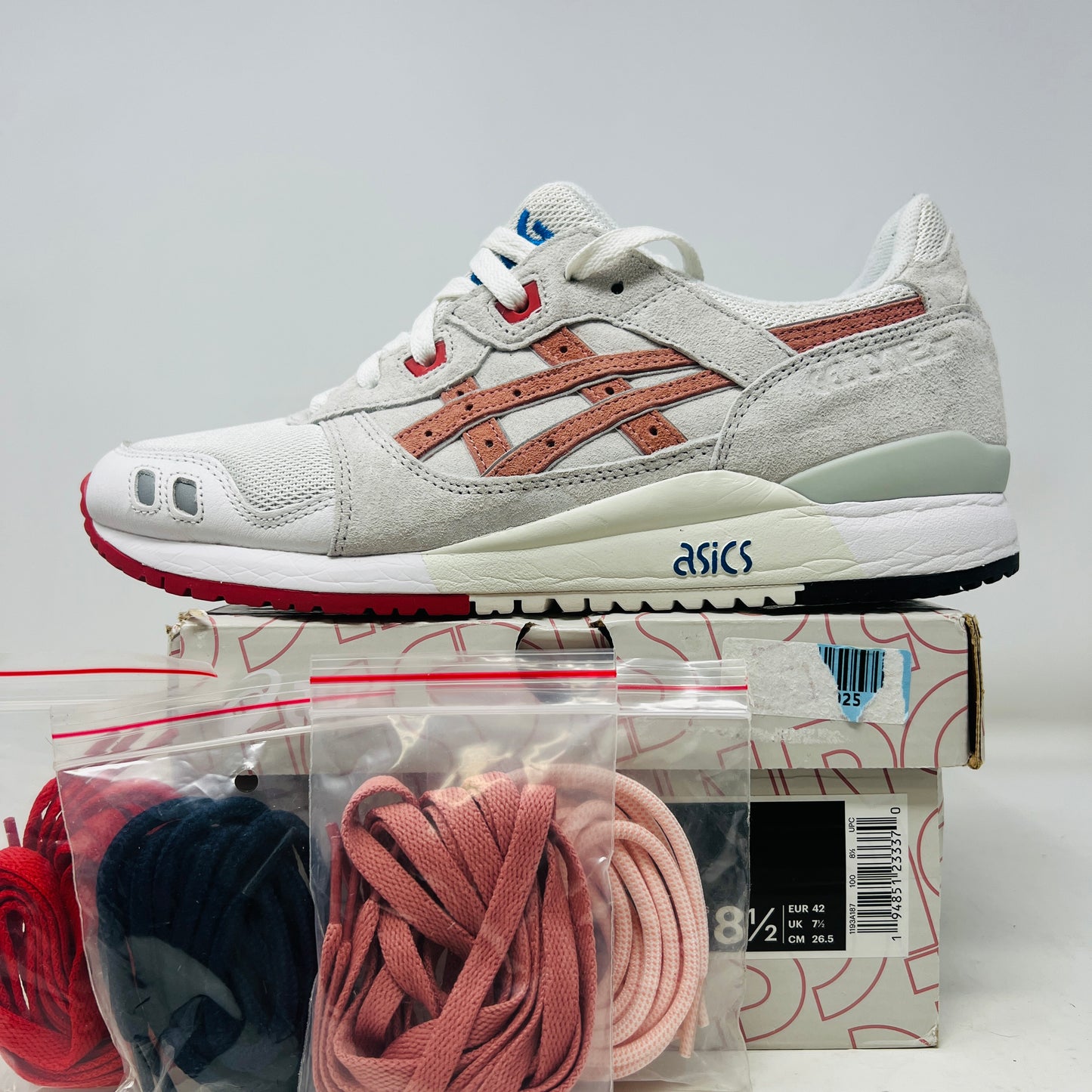 The ASICS Gel-Lyte III Ronnie Fieg Tokyo Trio Yoshino Rose sneaker, featuring clean white and beige uppers with red accents and pink iconic stripes, sits on its box. It includes navy, pink, and beige extra laces, with the authentic size clearly displayed on the box.