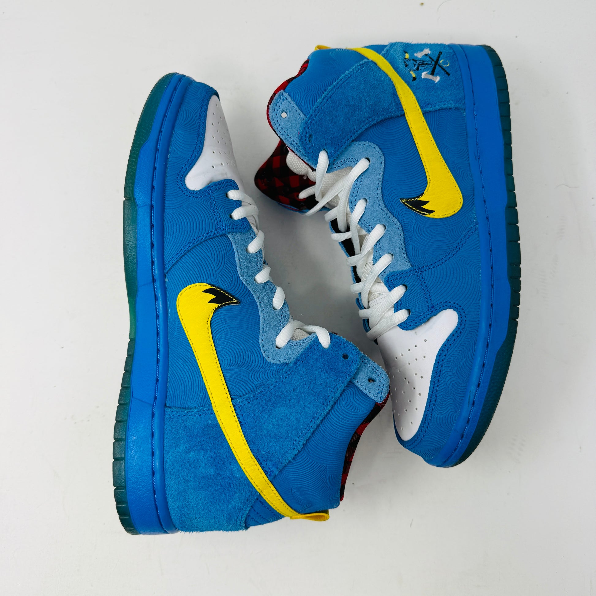 The Nike SB Dunk High Familia Blue Ox, a 2015 release by Nike, features clean blue uppers with a yellow swoosh, decorative stitching, red and black checkered lining, lace-up front, and an edgy skull with crossed bones on the side—its standout design loses no stars.