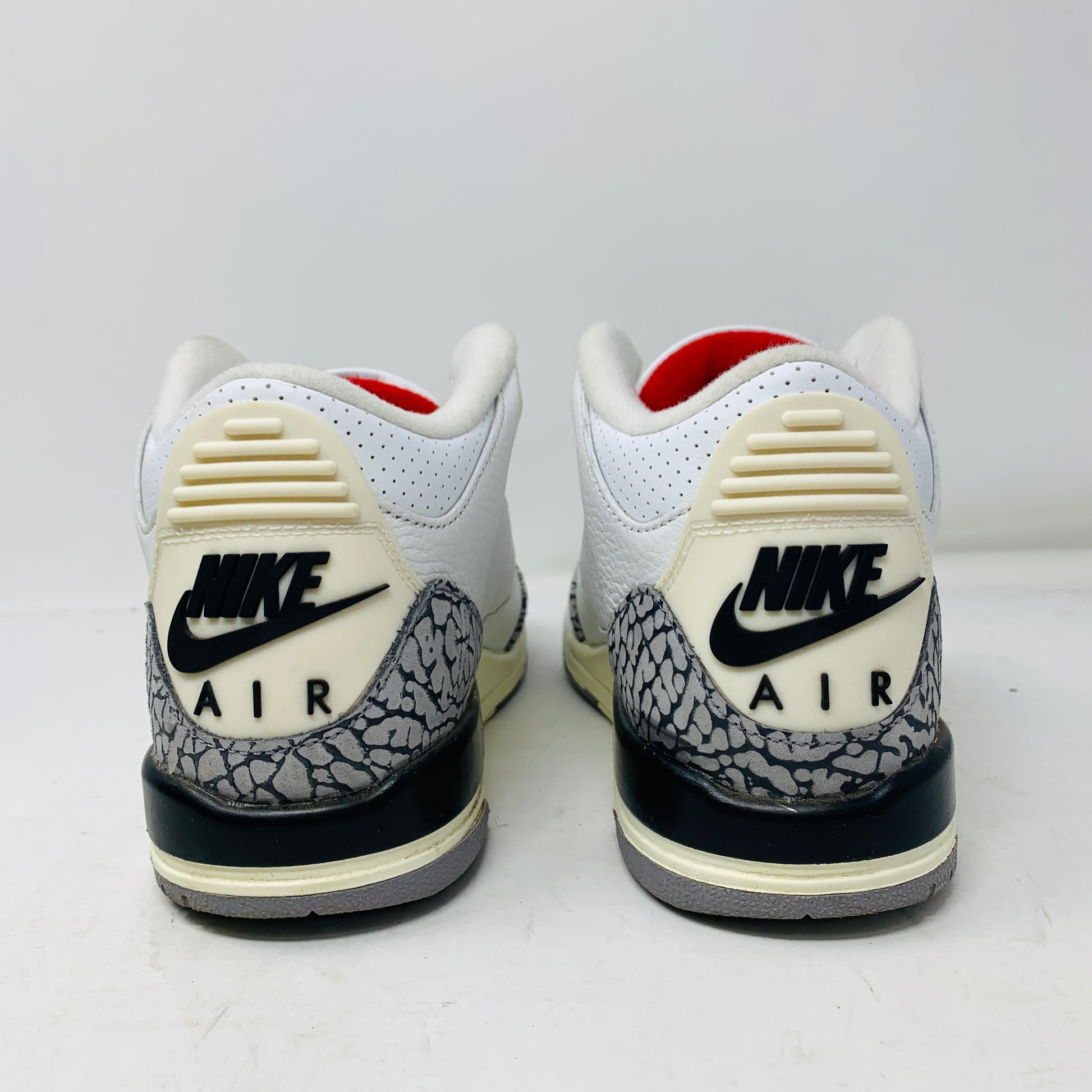 The image depicts the back view of a pair of Jordan 3 Retro White Cement Reimagined (GS) sneakers by Jordan, featuring white with black and gray elephant print accents, red lining, and a black Nike logo on the heel tab.