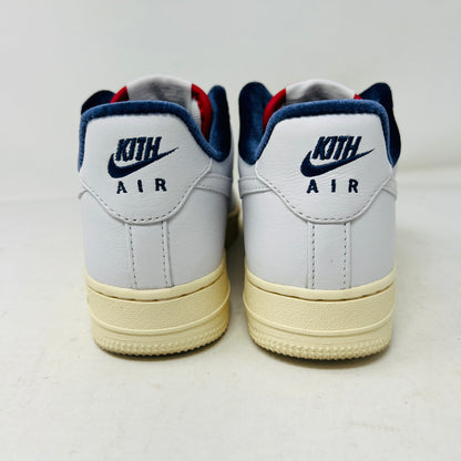 Rear view of Nike Air Force 1 Low Kith Paris sneakers shows KITH AIR in blue on heel, cream-colored soles, and dark blue trim. Background is a plain white surface.