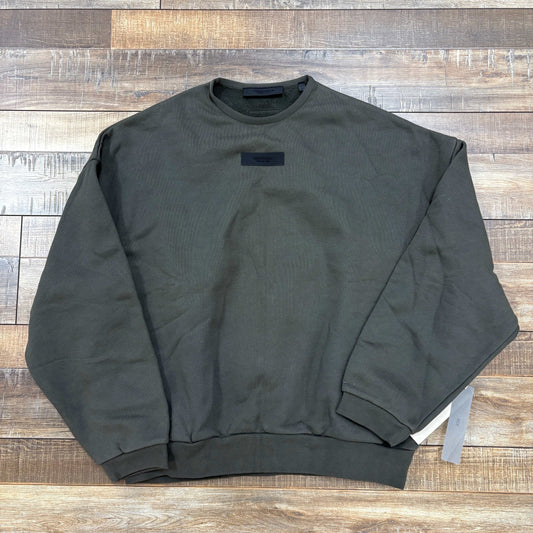 The Fear of God Essentials Crewneck Ink, an authentic dark green crewneck sweatshirt with long sleeves, lies flat on a wooden floor. A small label near the collar and ribbed cuffs and hem add appeal, while attached white tags at the side seam confirm its brand-new condition.