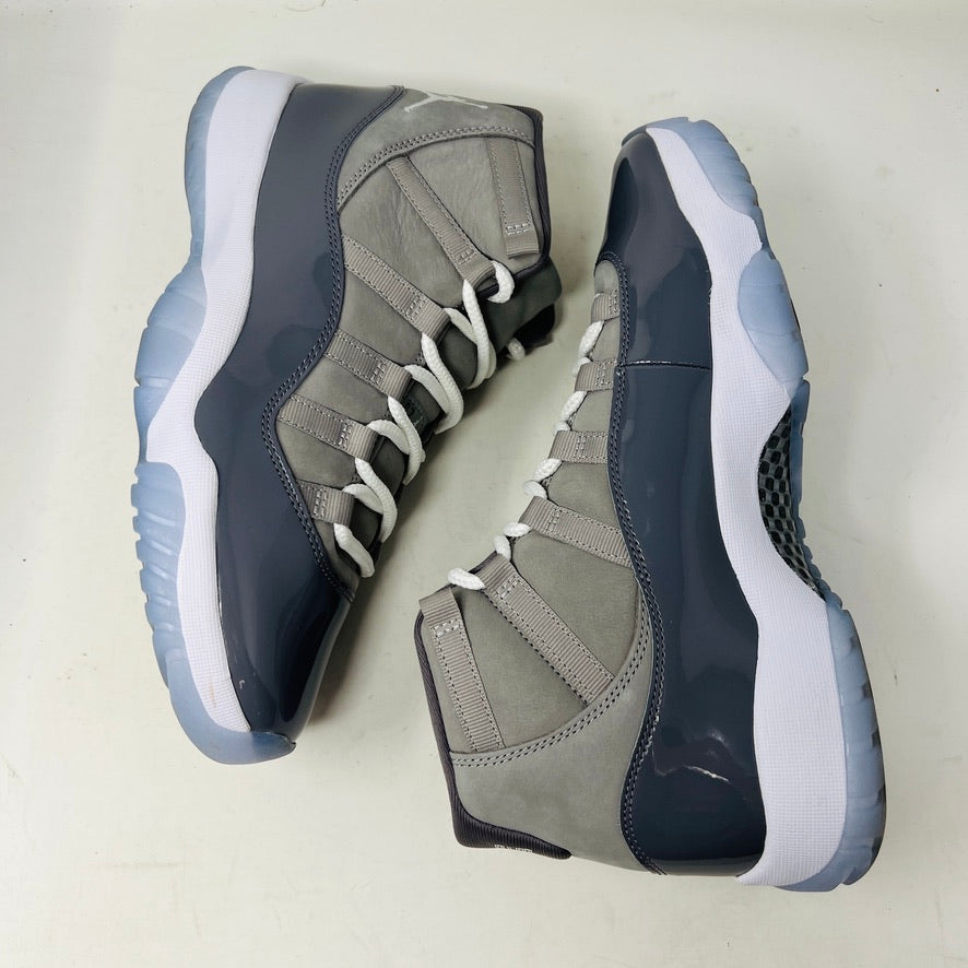 A pair of Jordan 11 Retro Cool Grey (2021) sneakers with white laces and translucent soles rest on a gray Nike Air box, showcasing the shiny patent leather and Jumpman logo. Size 9.5 appears on the label.