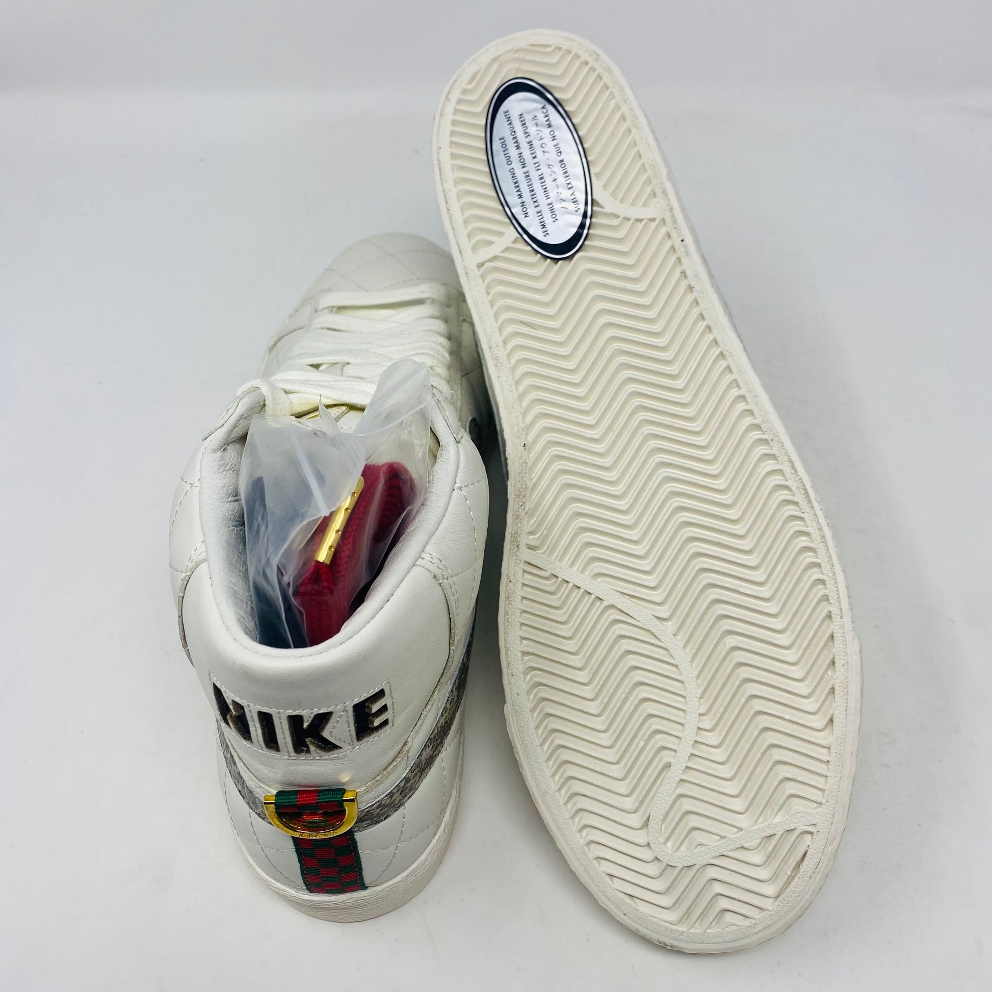 Nike SB Supreme Blazer White 2006 sneakers with embroidered text and a patterned emblem on the heel. The left shoe is upright with packaging inside, while the right shoe displays a grooved sole and an oval information label.