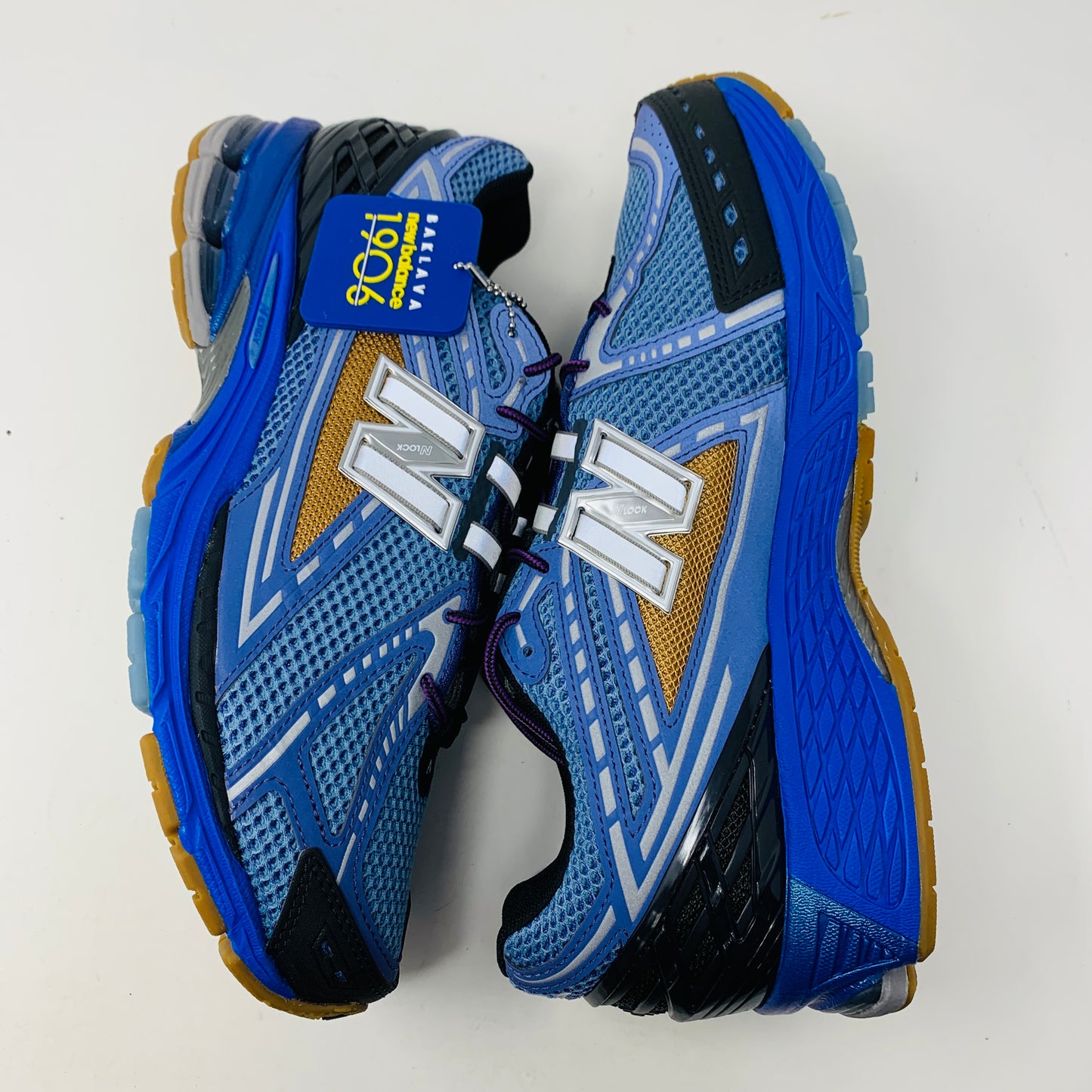 A brand new New Balance 1906R Action Bronson Medusa Azul sneaker with intricate designs and a silver N logo rests atop an orange box labeled BAKLAVA new balance. The 2024 edition features a layered design with black and metallic accents, complete with an intricate sole and dust bag.