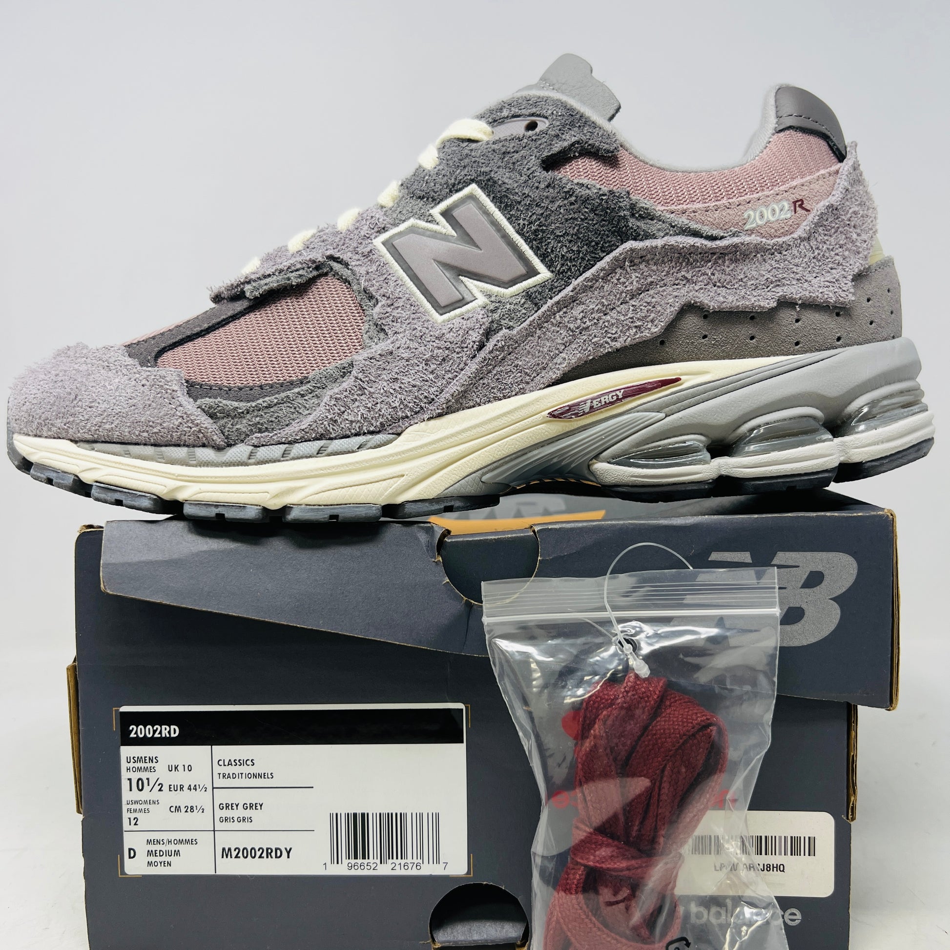 A pair of New Balance 2002R Protection Pack Lunar New Year Dusty Lilac sneakers mix gray and beige tones with CLEAN UPPERS and a textured design. Displayed on a shoebox, they include extra maroon laces. The box label details the shoe size and model.