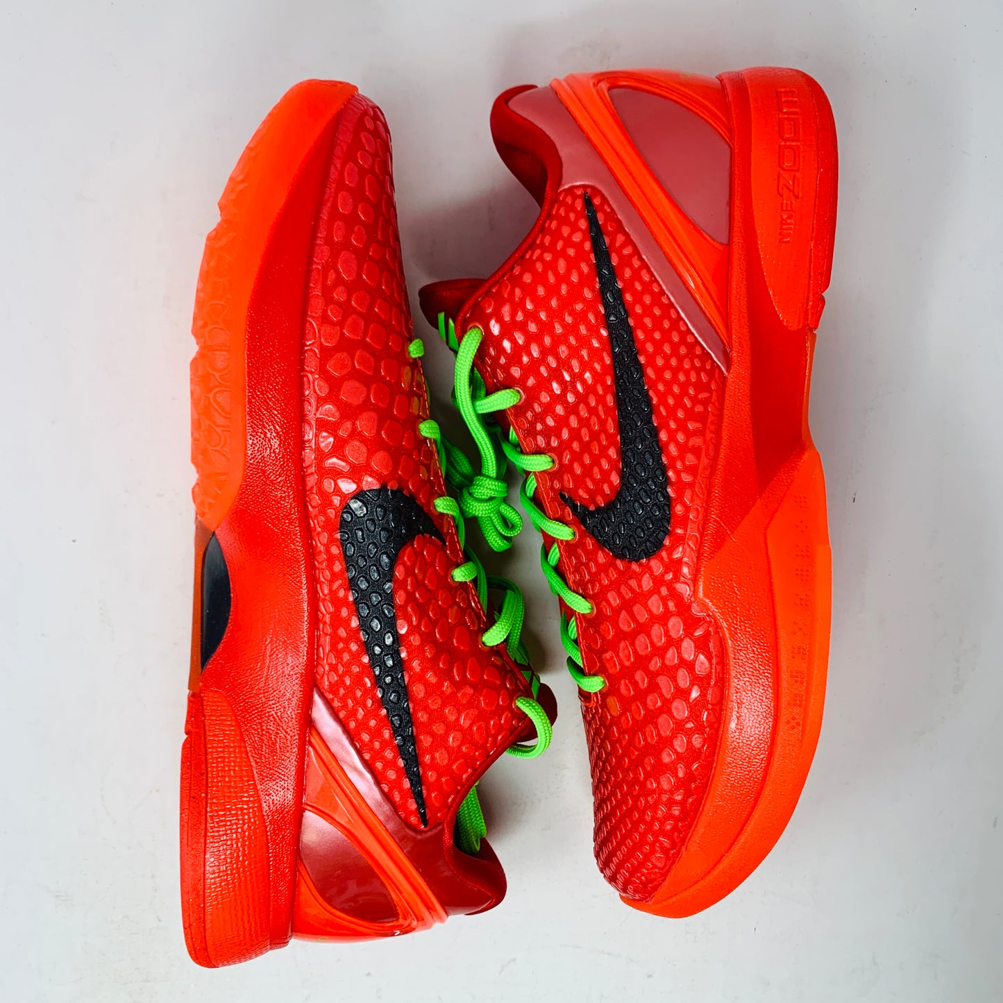 The Nike Kobe 6 Protro Reverse Grinch basketball sneakers, featuring a textured red design, black swooshes, and vibrant green laces, are placed side by side against a light background.