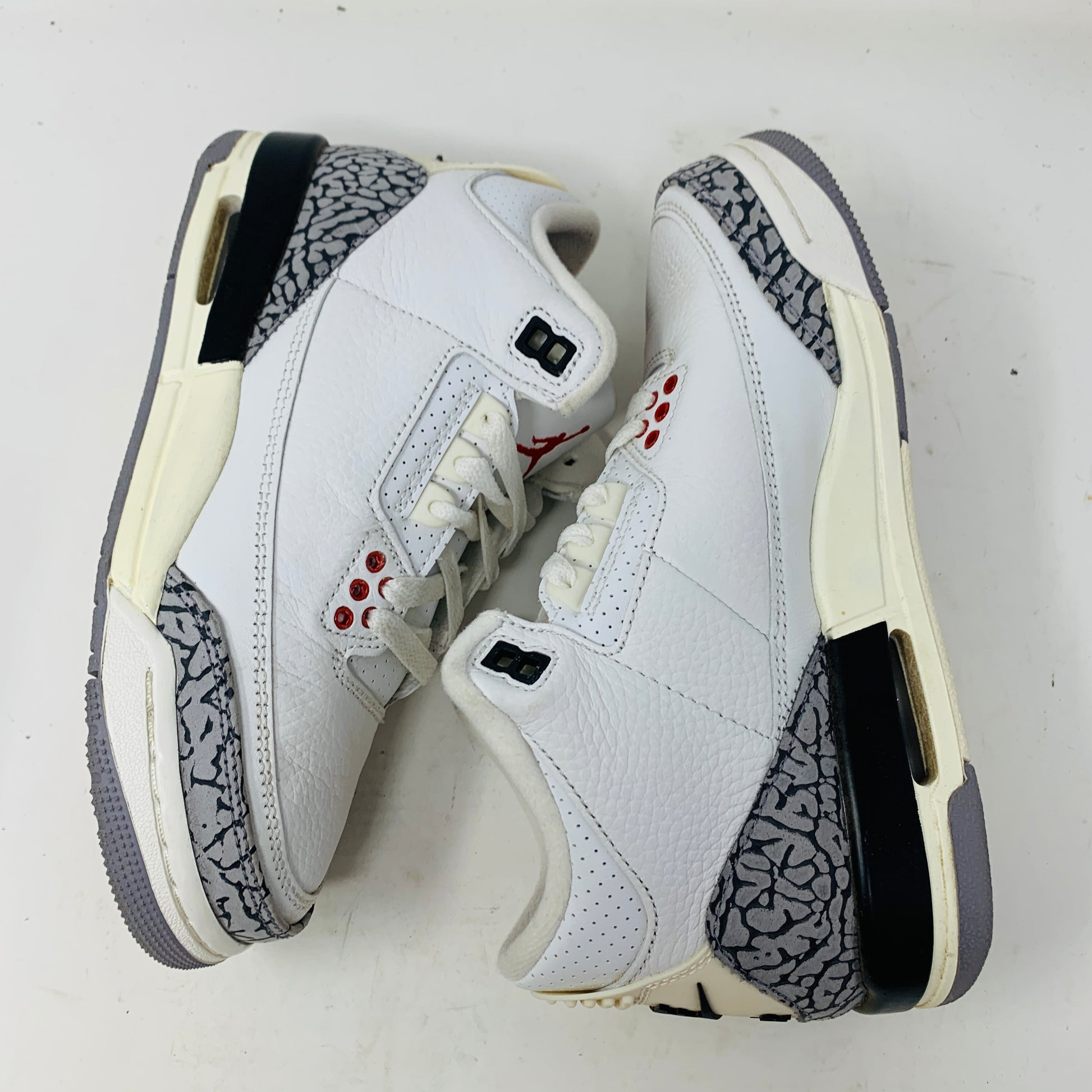 The Jordan 3 Retro White Cement Reimagined (GS) sneakers, branded by Jordan, are white with gray and black accents, red eyelets, and elephant print details on the sides and heel. They are displayed side by side on a white surface.