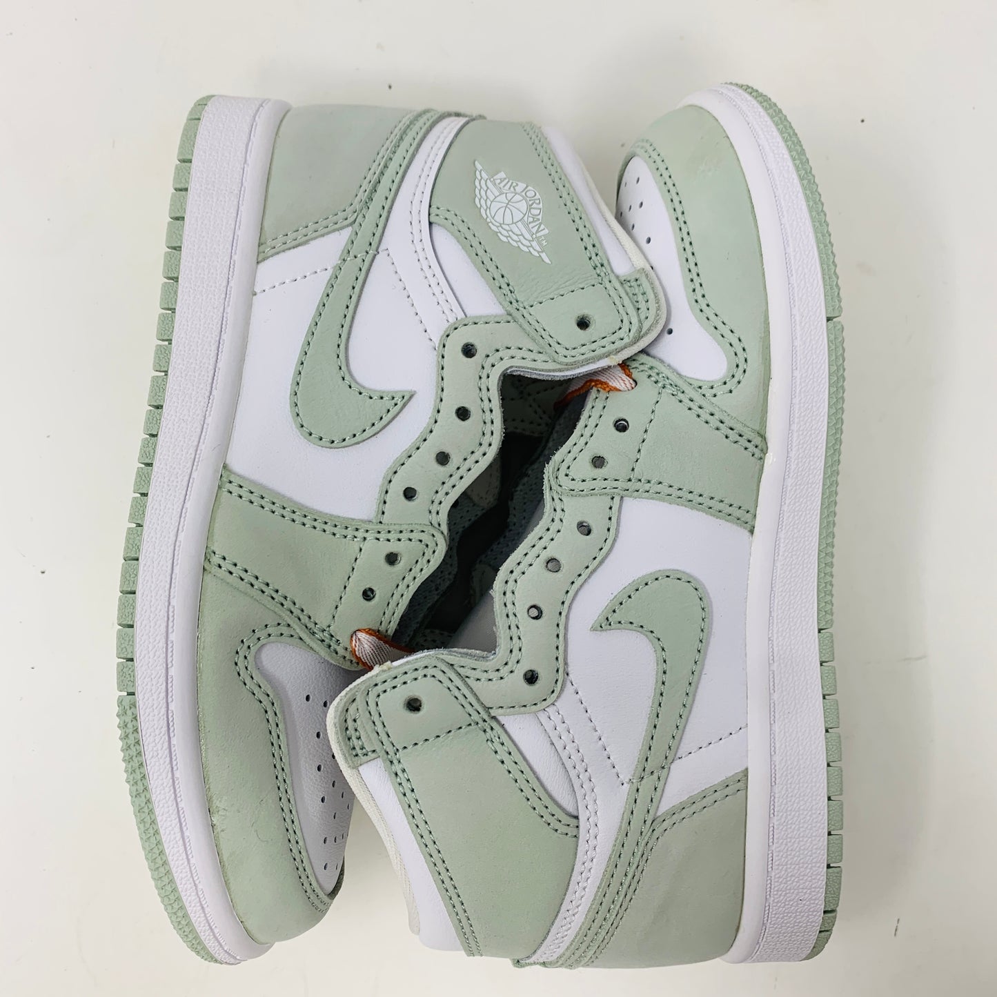 The Jordan 1 Retro High OG Seafoam (PS) by Jordan are high-top sneakers in a white and light green color scheme, featuring a wing logo and swoosh on the sides, positioned sole to sole on a plain white background.