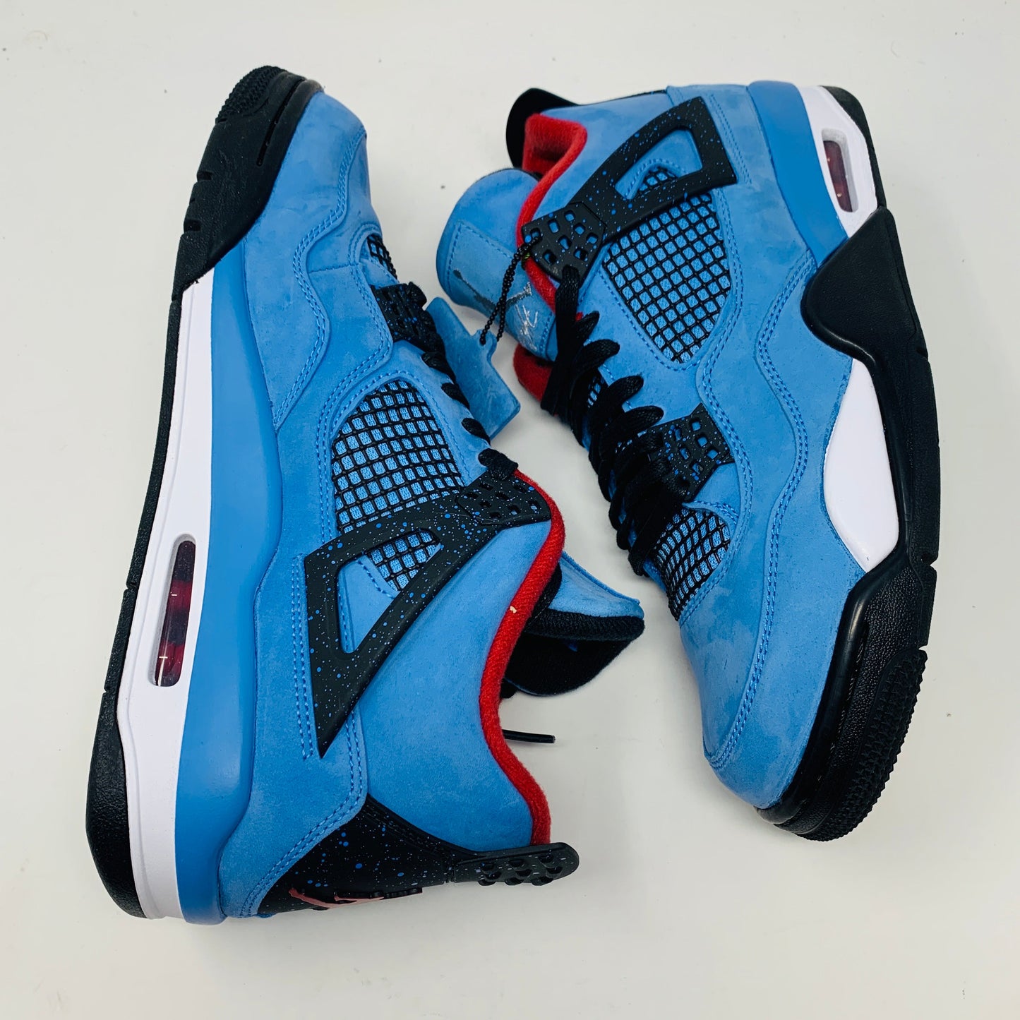 The Jordan 4 Retro Travis Scott Cactus Jack sneaker in blue, accented with black and red, features a visible air unit in the heel and a Jumpman logo hang tag, packaged in a black speckled box with an orange Jumpman logo and AIR.