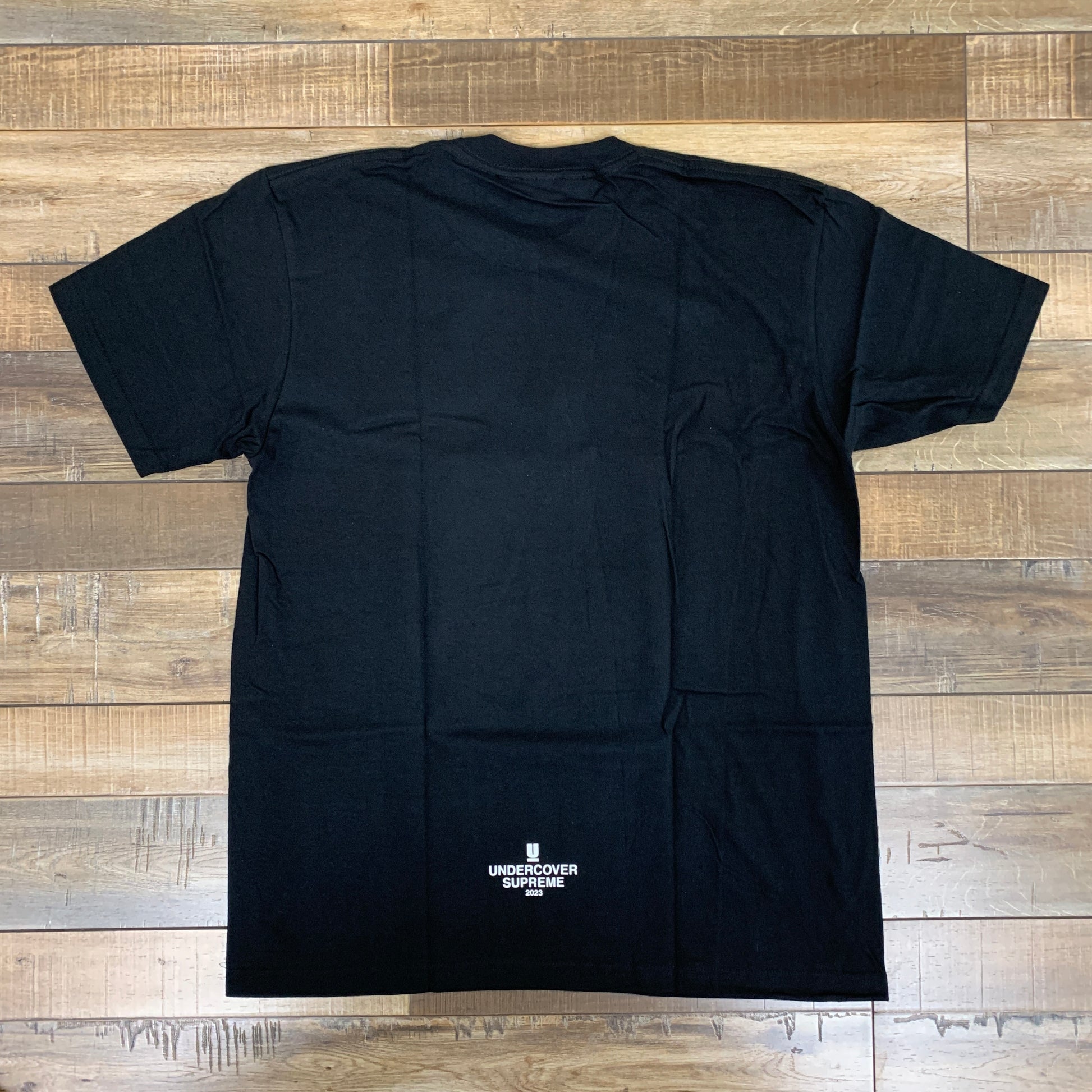 The Supreme UNDERCOVER Face Tee in black, featuring a small white logo and text near the bottom, is displayed on a wooden floor.