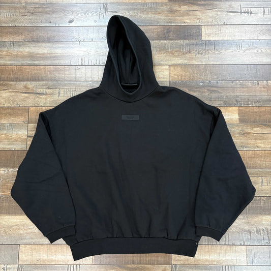 The Fear of God Essentials Chest Logo Hoodie in Jet Black, a 100% authentic piece from Essentials, is laid flat on a wooden floor. Its hood rests neatly on the back, showcasing its stylish front pocket and long sleeves—an essential blend of style and comfort.